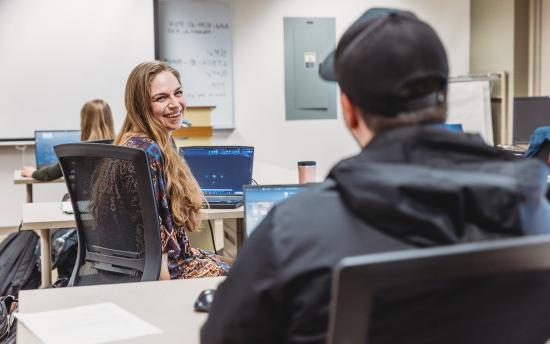 A student looking over her shoulder smiling at the camera with a laptop open in front of her